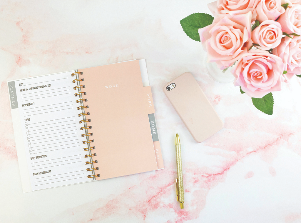 A peach-colored planner, a gold ballpoint pen, a smart phone with a peach-colored case, and a bouquet of pink roses on a pink and white marbled surface