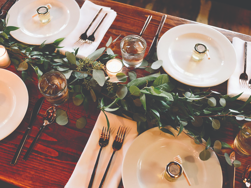 Place settings on a greenery-lined wooded table, including white plates, elegant flatware, and individual jars of honey as wedding favors