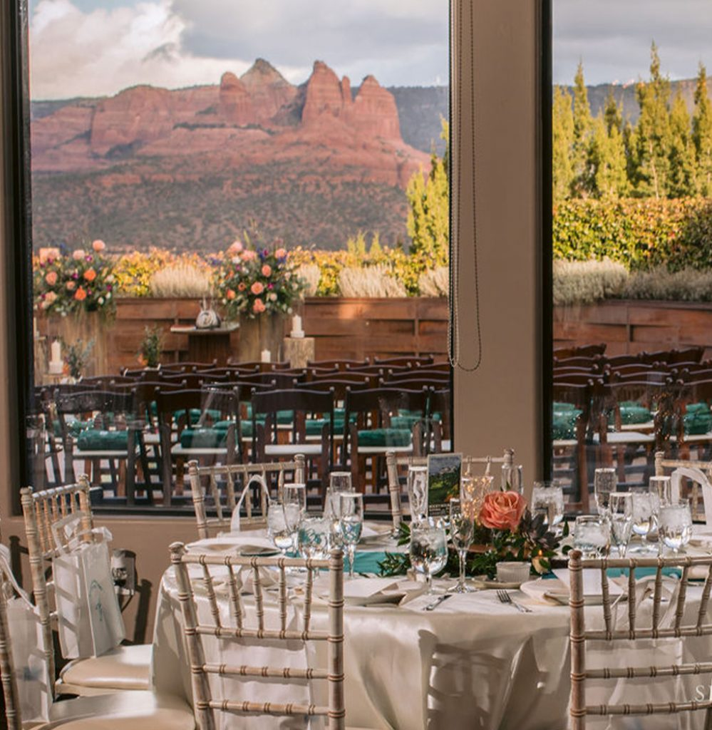 A classy dining hall at Agave of Sedona — Wedding and Event Center with light-colored chairs and table settings in the foreground and dark-colored chairs and a Sedona landscape are visible through large windows in the background
