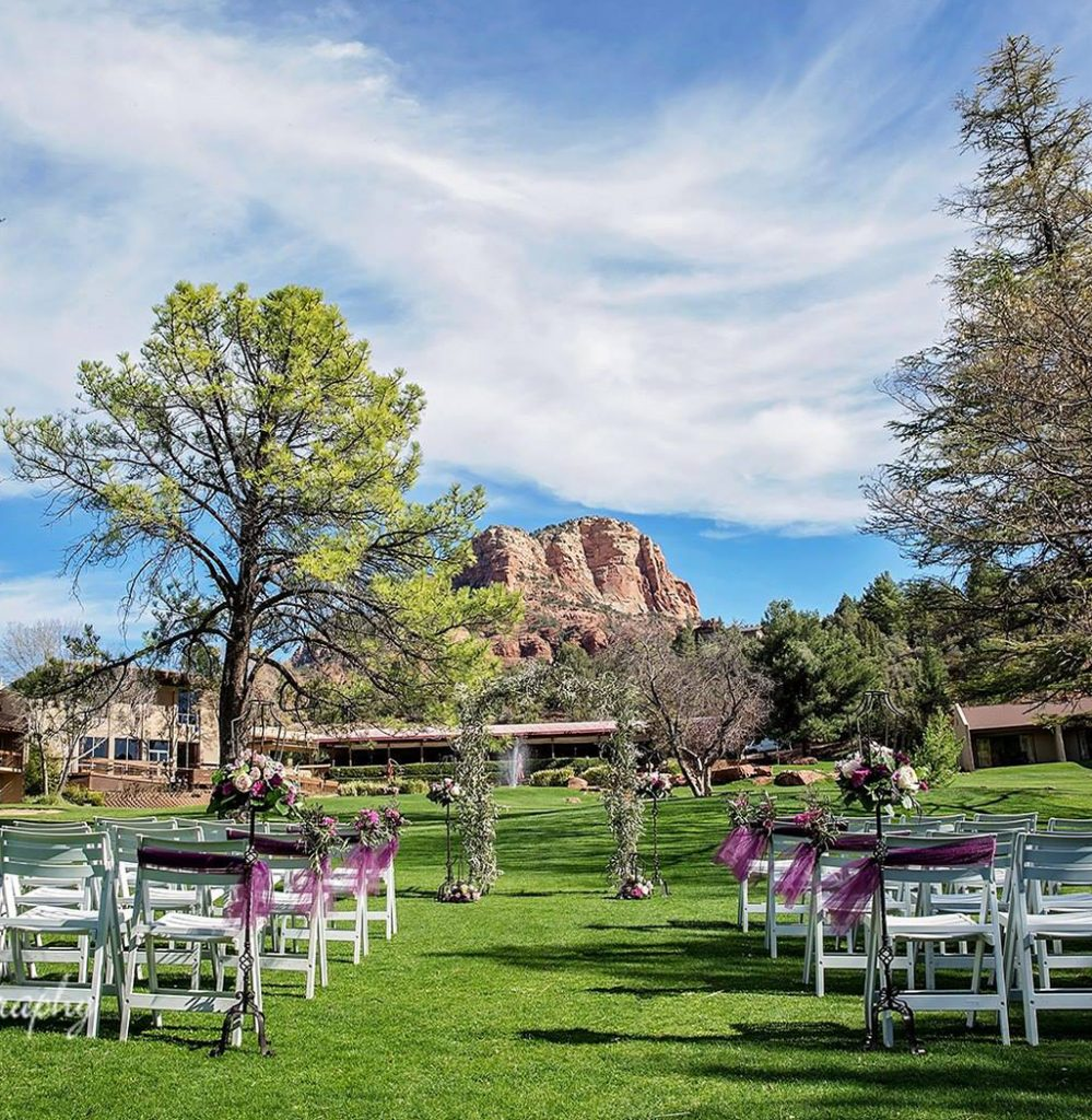 White chairs, some adorned with purple tulle, sit in rows on either side of a grassy wedding aisle with a flowery arch outdoors with Sedona’s Red Rock formations in the background at Poco Diablo Resort