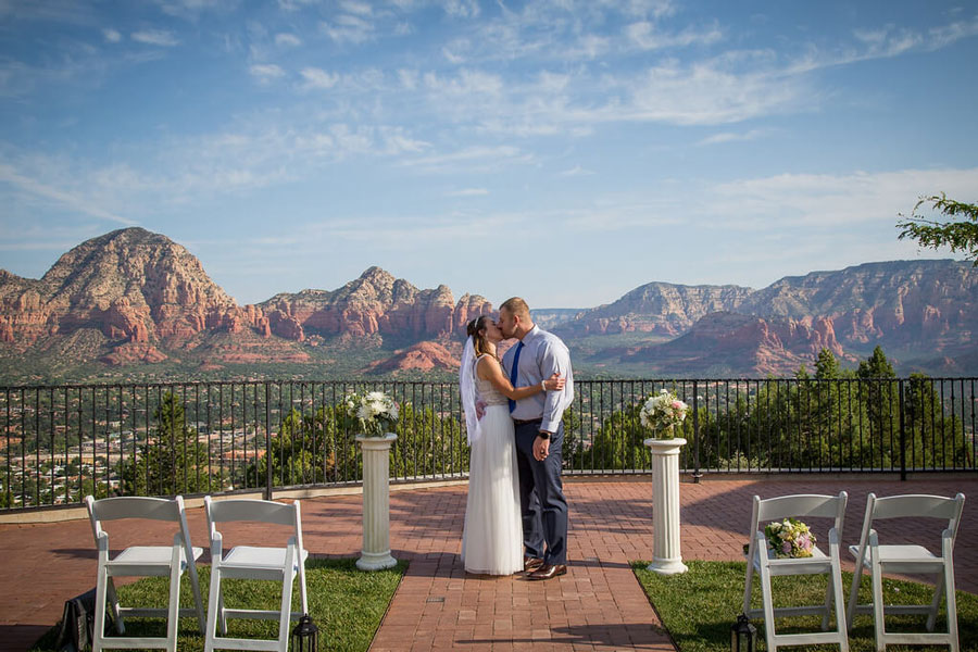 A bride and groom kiss between two small white pillars with the rocky landscape near Sedona in the background