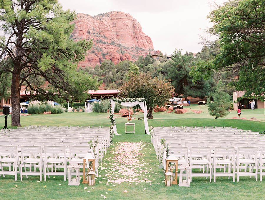 An outdoor wedding setup with white chairs, an arch adorned with fabric and flowers, flower petals lining the aisle, and Red Rock formations in the distance