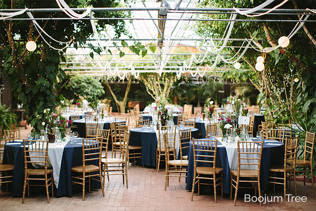 Natural woden chairs arranged in around tables covered with navy-colored tablecloths with white runners and simple floral centerpieces adorn a charming brick foundation patio with greenery and lights all around