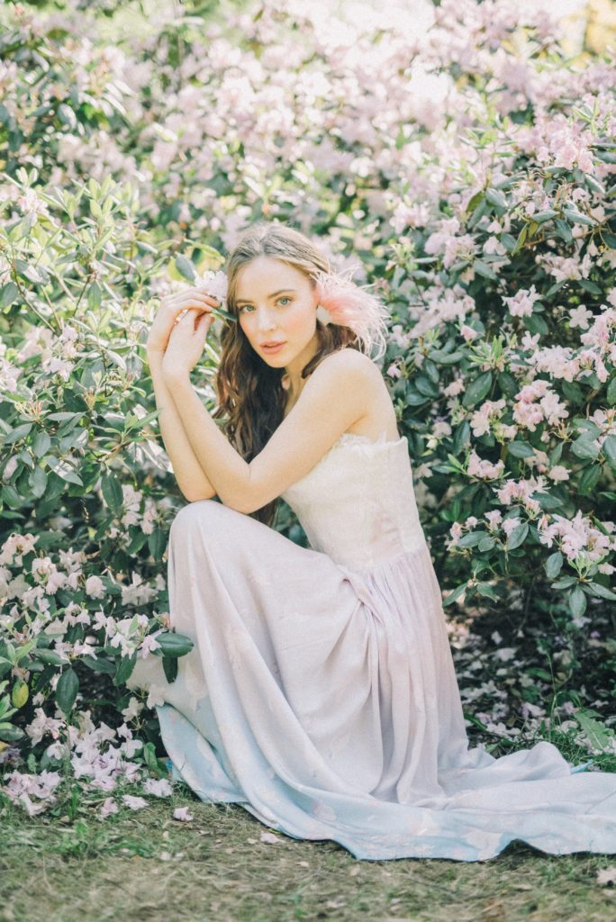 a bride in a long dress crouches next to a bush with light pink flowers