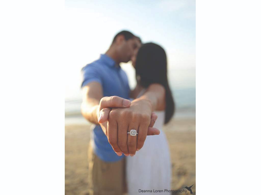 A couple holds hands to show off a square engagement ring on the bride-to-be’s finger in the foreground while the couple kissing is out of focus in the background