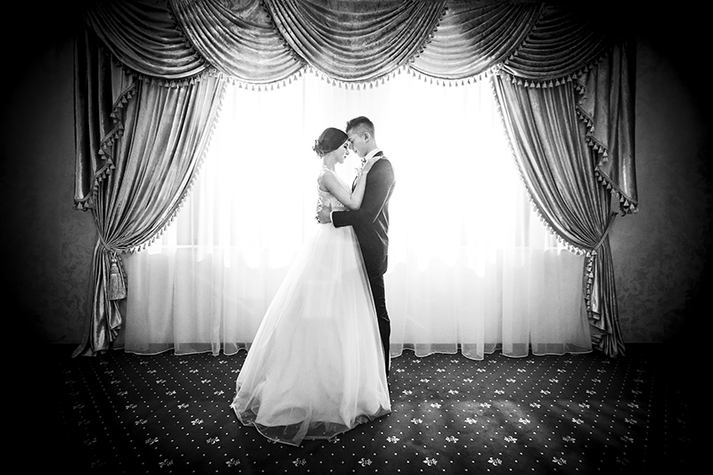 Newlywed couple standing in front of a resort window. Image is black and white and dramatic. Great wedding photography example.