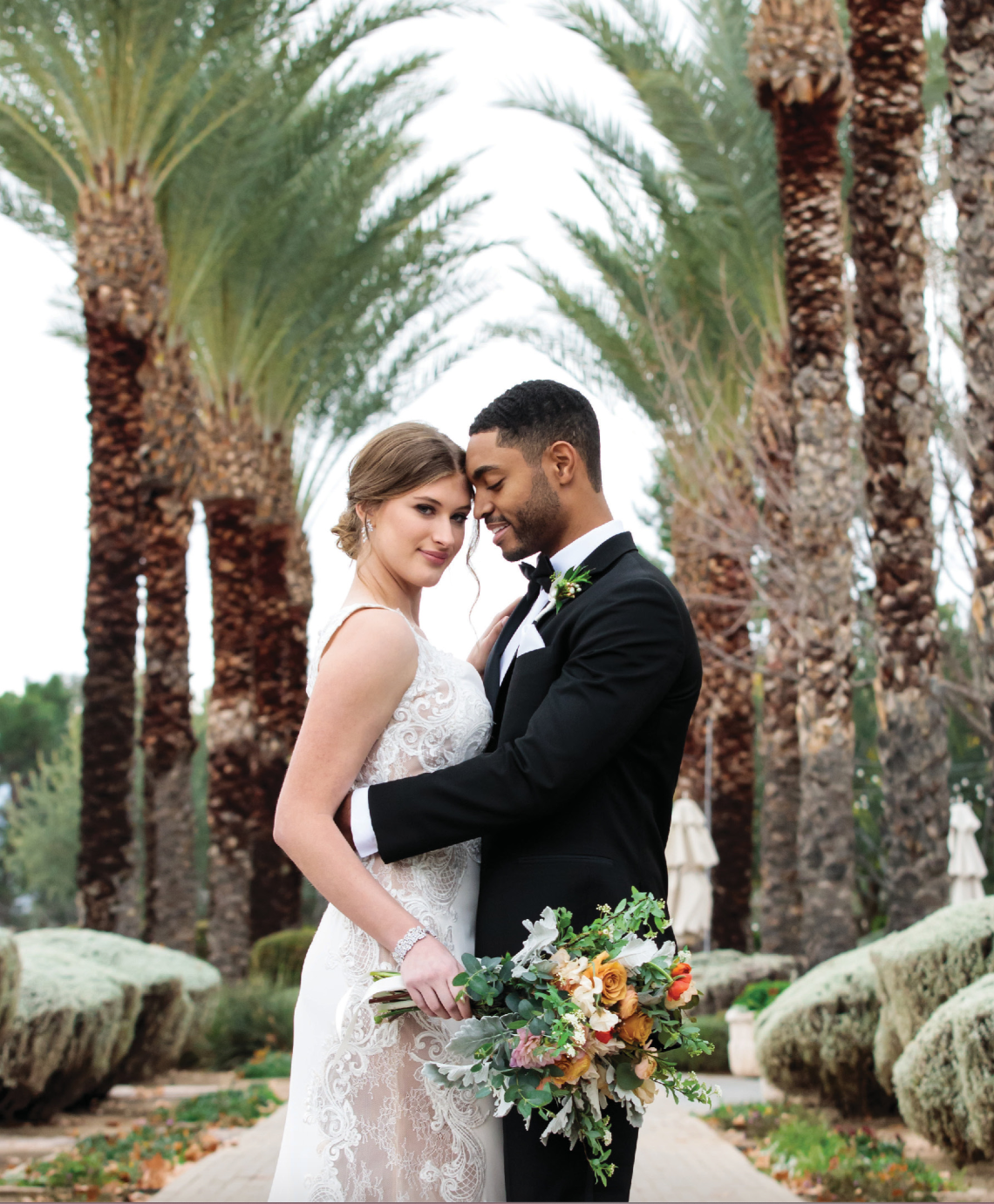 Bride holding floral bouquet and embracing groom in elegant black tux on a walkway lined with tall palm trees at a Tucson Wedding venue. 