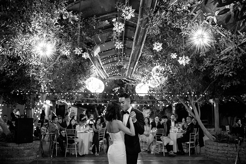 Classic black and white picture of wedding couple dancing at their catered dinner in an outdoor garden covered with string lights and lanterns in the trees. 
